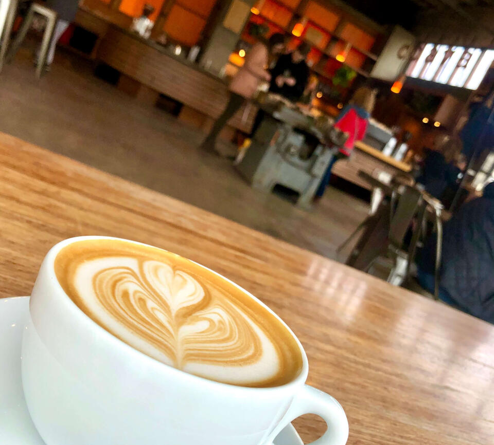 A coffee cup with latte art sits on a wooden table in an airy cafe